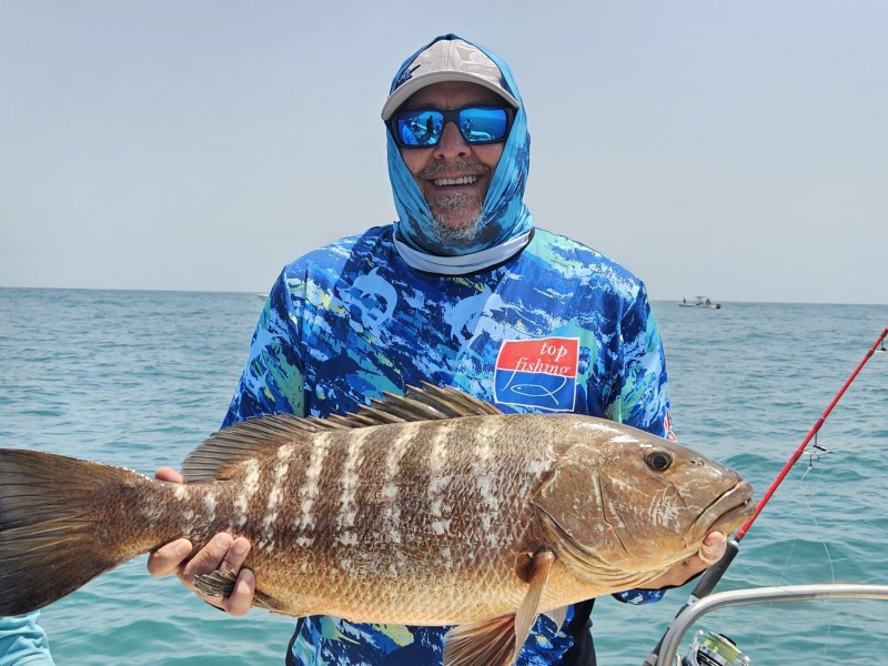 Un pêcheur souriant pose en mer avec une belle prise : une carpe rouge (ou mangrove snapper) aux flancs zébrés.